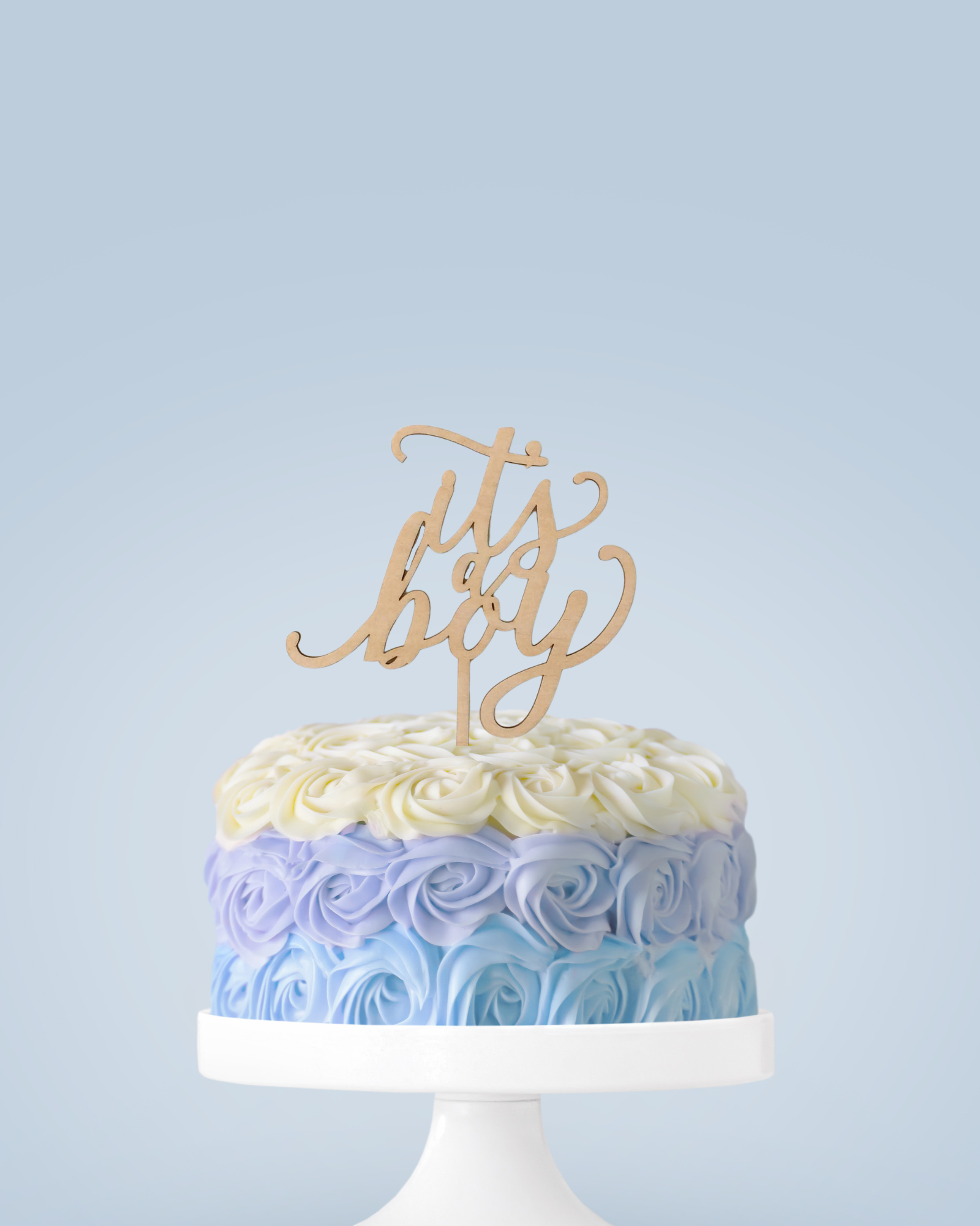 A layered cake with blue and white rose decorations and a wooden topper reading "it's a boy" on a white stand.