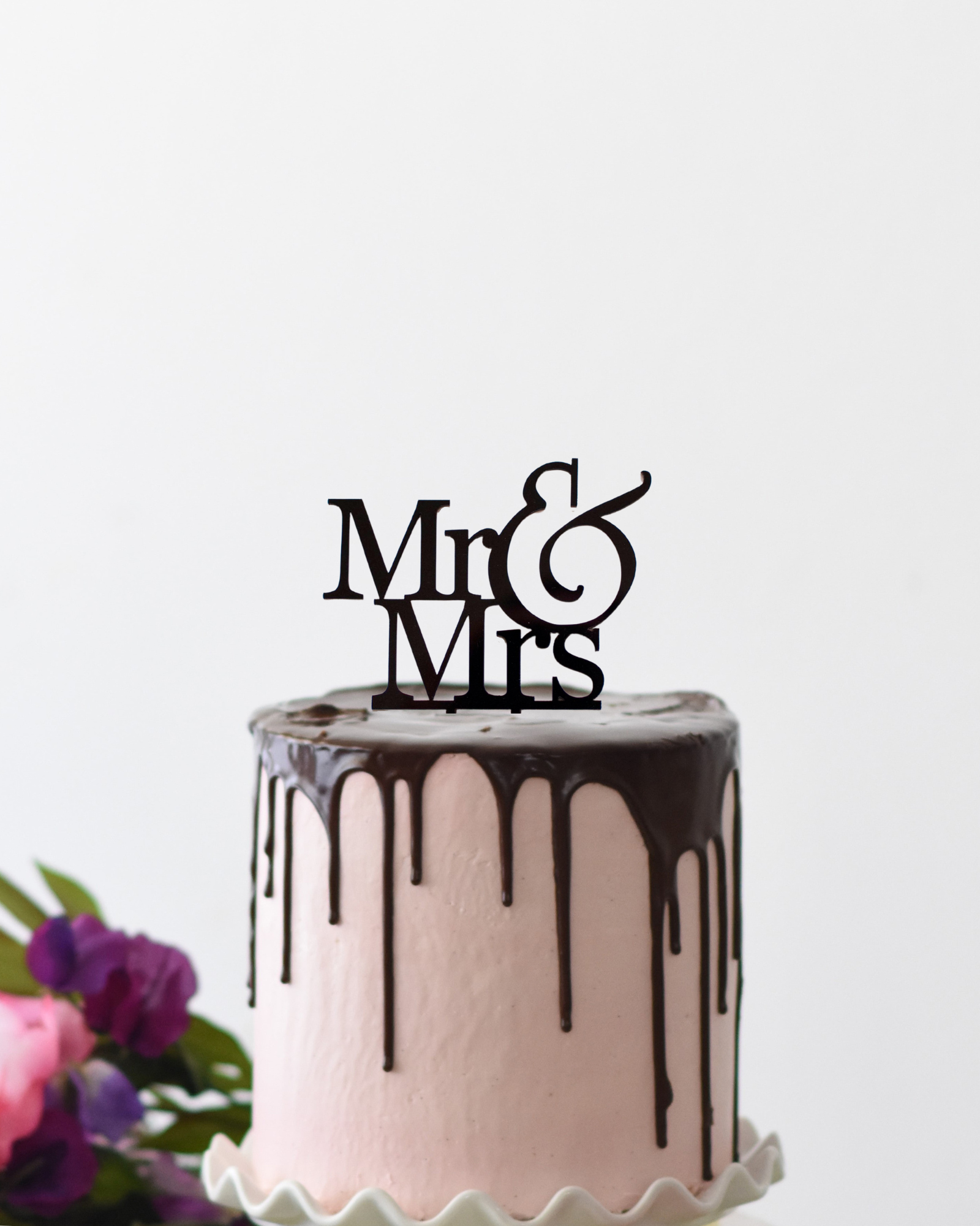 A pink frosted cake topped with a black "Mr & Mrs" topper, surrounded by vibrant flowers.