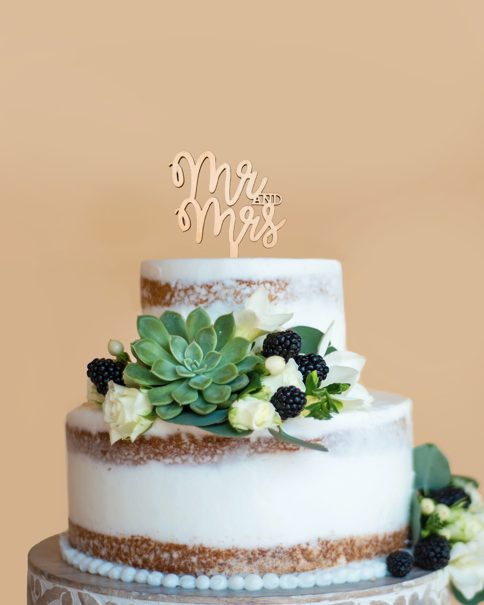 Two-tiered cake adorned with fresh flowers, berries, and a "Mr and Mrs" topper against a soft beige background.