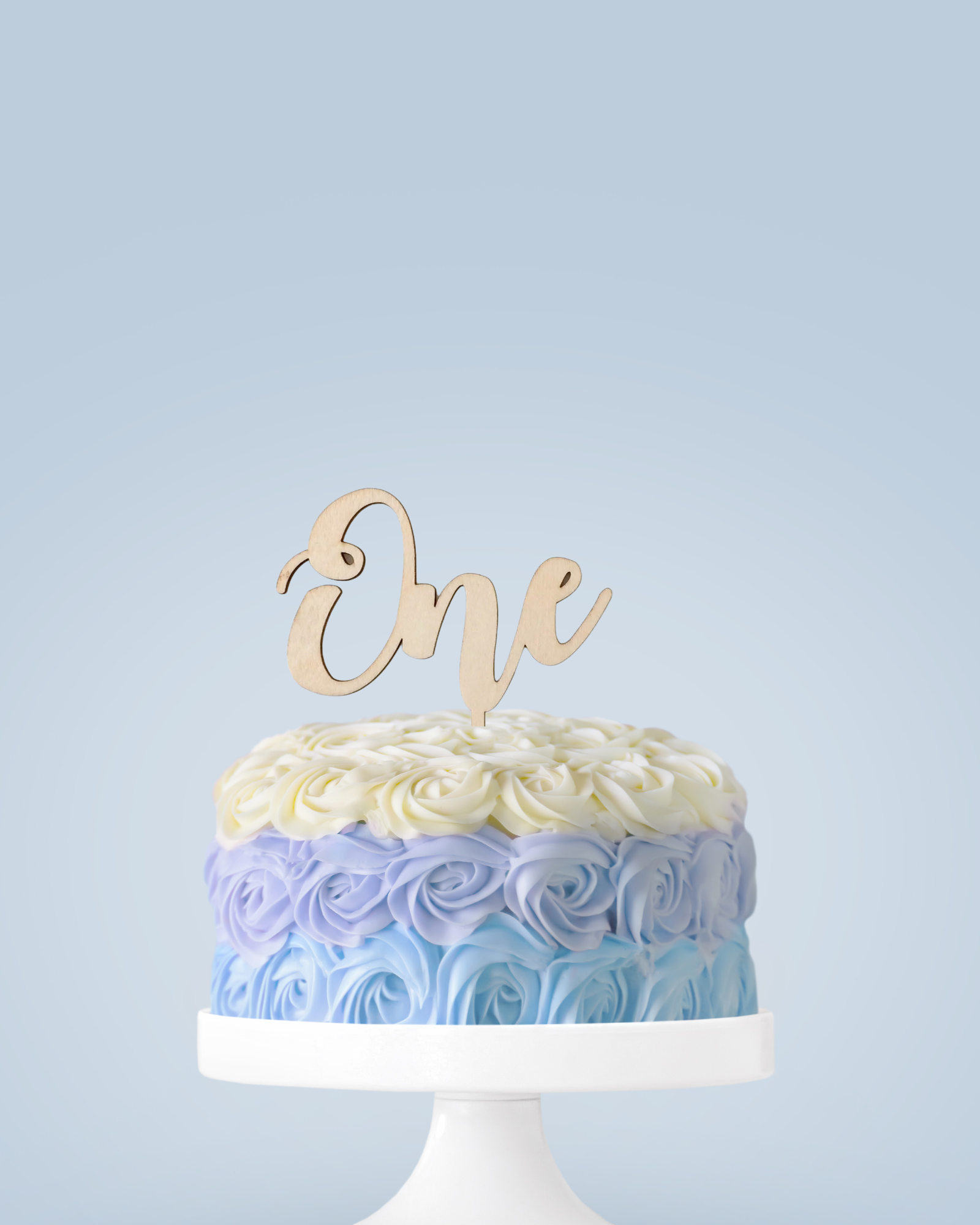 A beautifully decorated cake with pastel blue and yellow rosettes, topped with a wooden "One" sign.