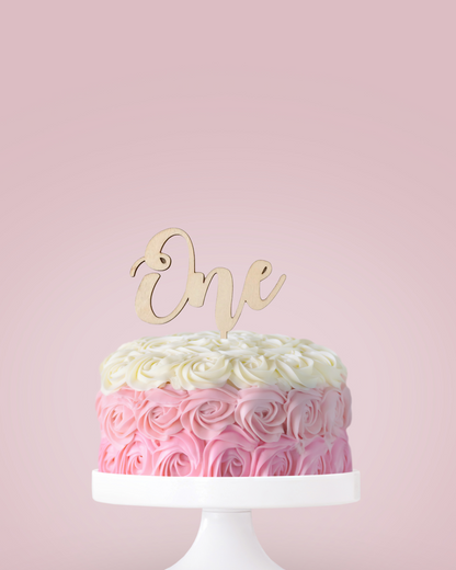 A pastel pink cake adorned with rose swirls, topped with a wooden "One" decoration against a soft pink backdrop.