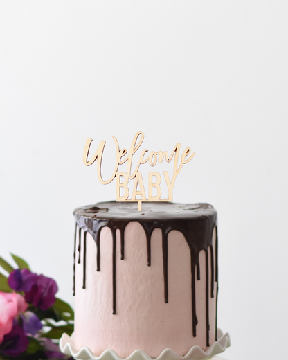 A pink cake with chocolate ganache, topped with a wooden "Welcome Baby" topper, surrounded by colorful flowers.