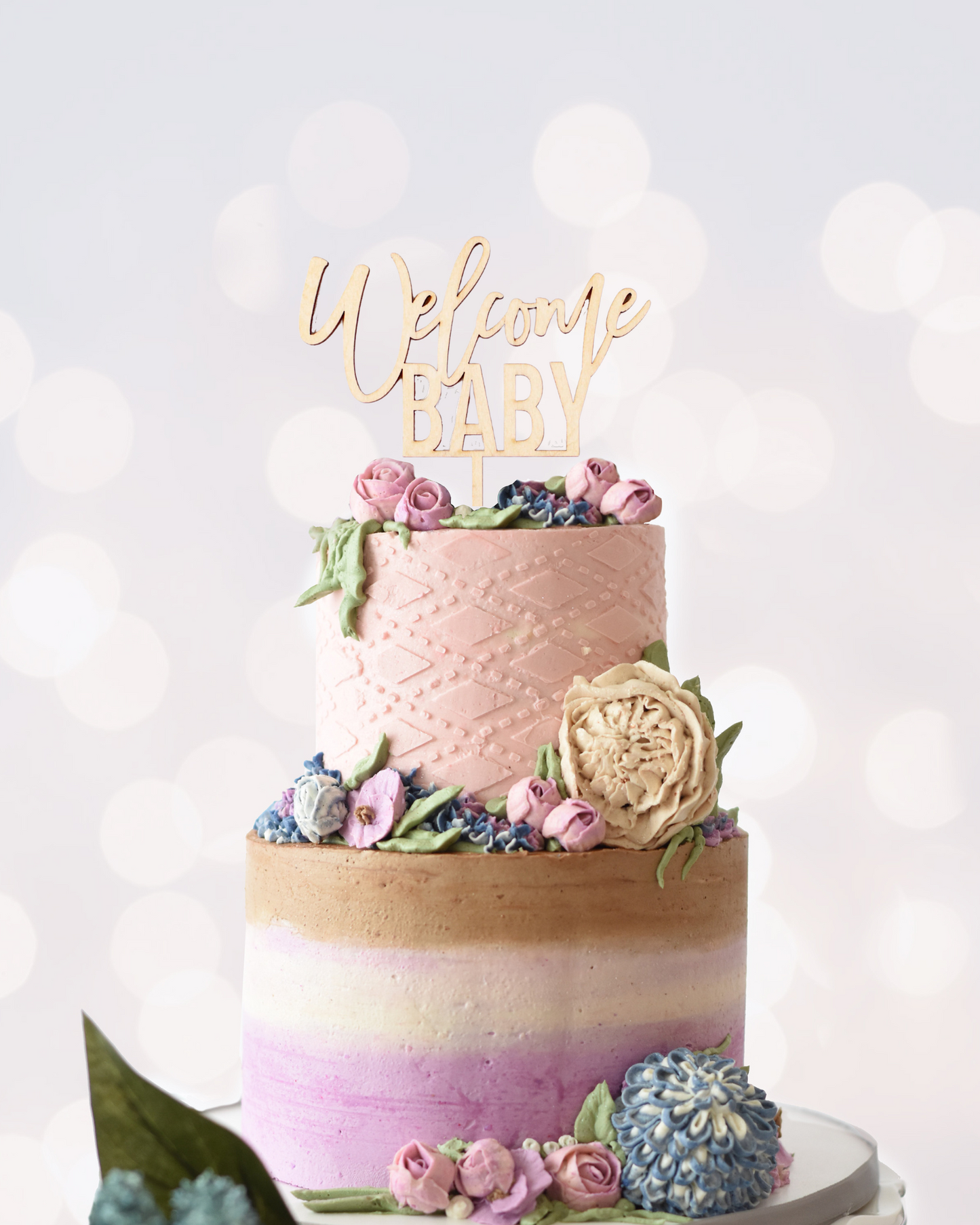 A tiered cake with pastel icing, floral decorations, and a "Welcome Baby" topper against a soft, blurred background.