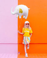 Girl in bright clothes, leaning against orange and pink wall holding a while foil elephant balloon