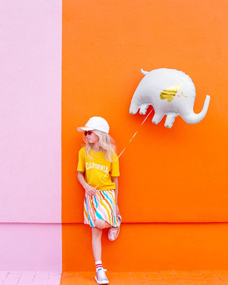 Girl in bright clothes, playing with shoe and looking to her right while leaning against orange and pink wall holding a while foil elephant balloon