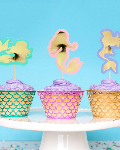 Three whimsical cupcakes with purple frosting topped with colorful mermaid silhouettes against a bright blue background.