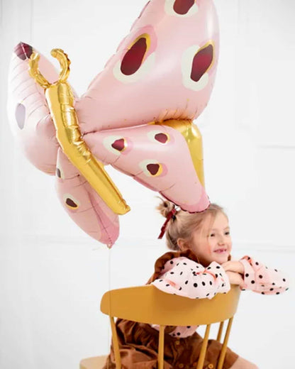 A child with a cheerful expression leans on a bright yellow chair, accompanied by a large pink butterfly balloon.