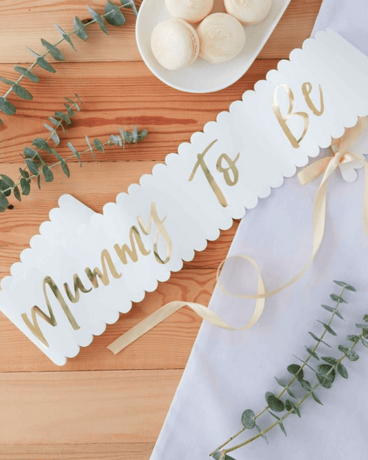 A white banner with "Mummy To Be" in gold script, surrounded by eucalyptus leaves and a plate of macarons.