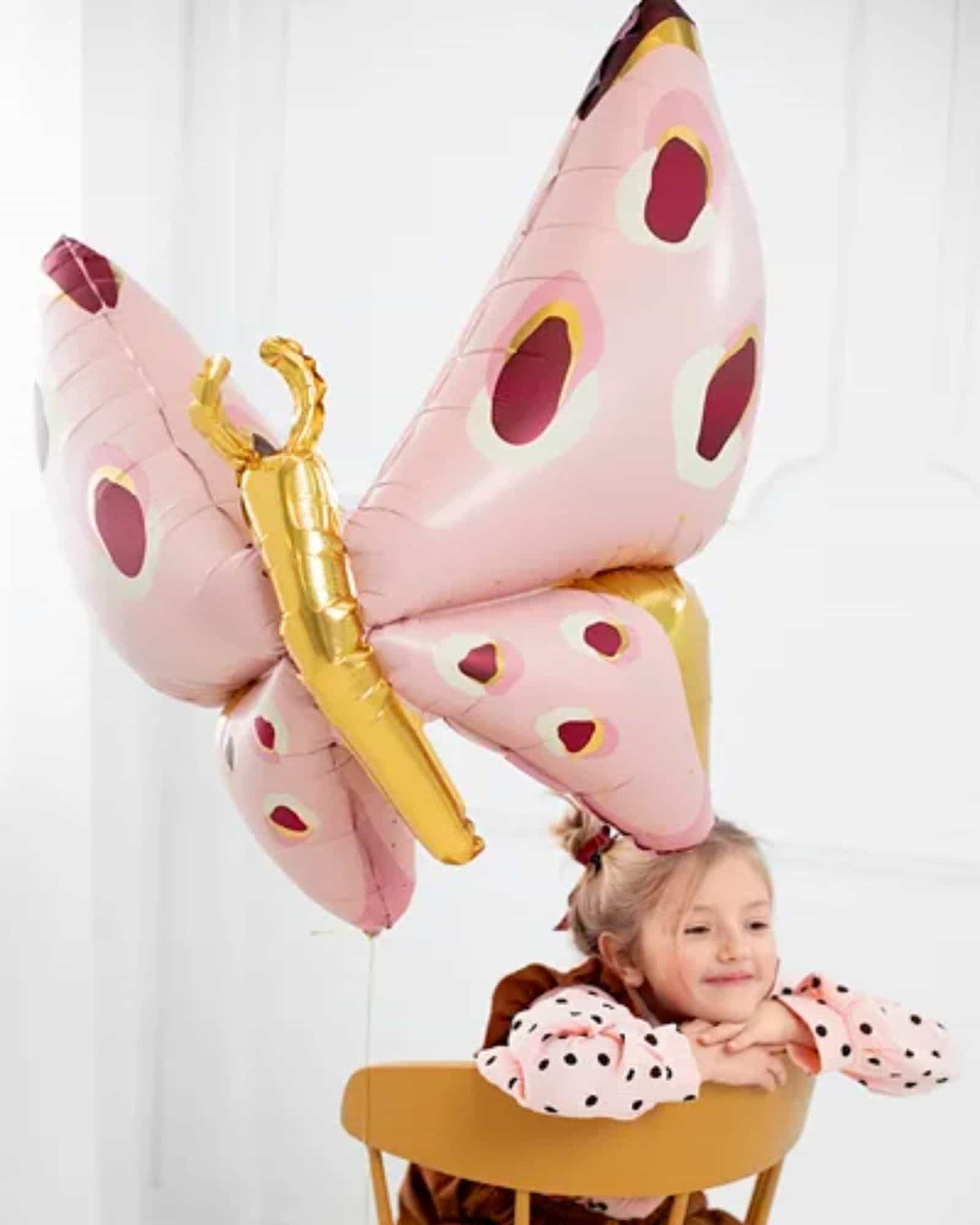 A child with a playful expression rests on a chair, adorned with a large, colorful butterfly-shaped balloon overhead.