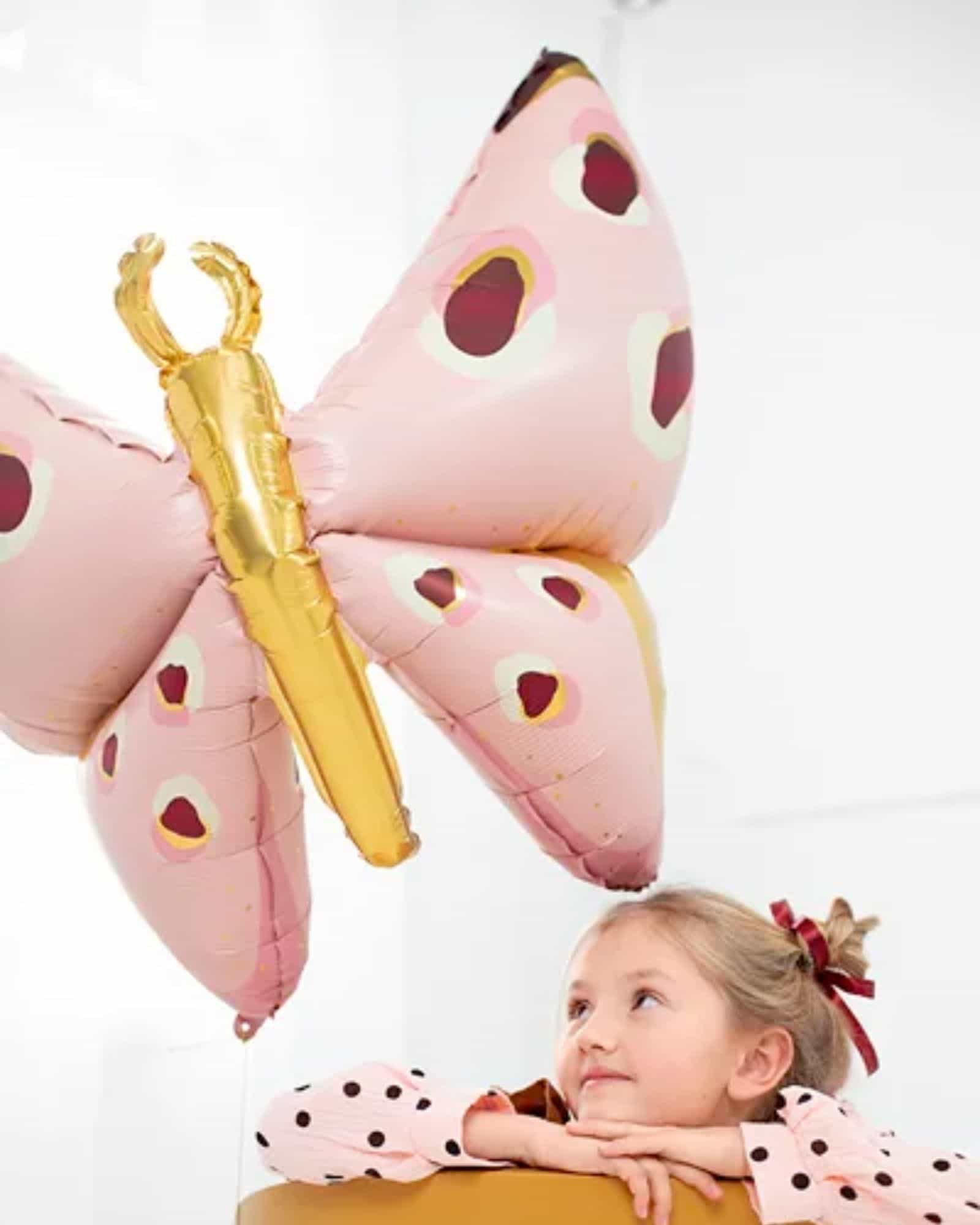 A girl with a polka dot shirt gazes up at a large pink butterfly balloon with gold accents.