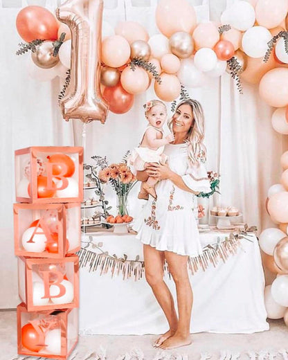 A woman holds a toddler in a festive setting with pastel balloons, floral arrangements, and a decorated table.