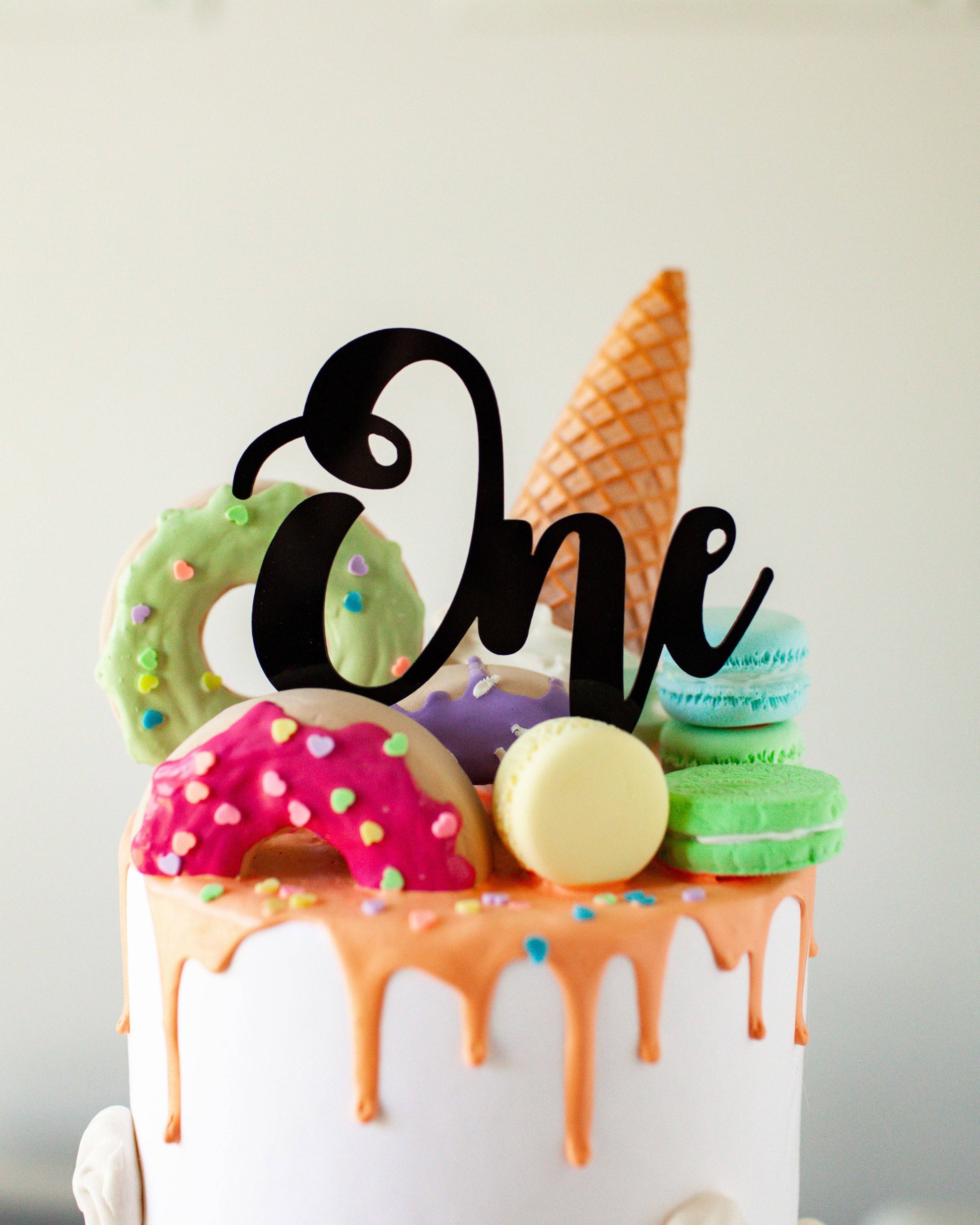 Black acrylic "One" topper atop a colorful cake adorned with donuts, macarons, and an ice cream cone.