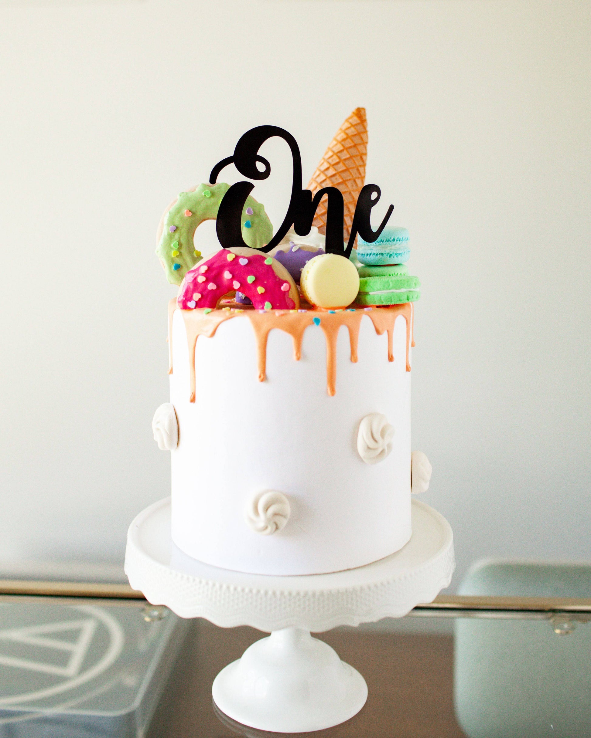 A white cake with colorful macarons, donuts, and an ice cream cone, topped with a black "One" sign.