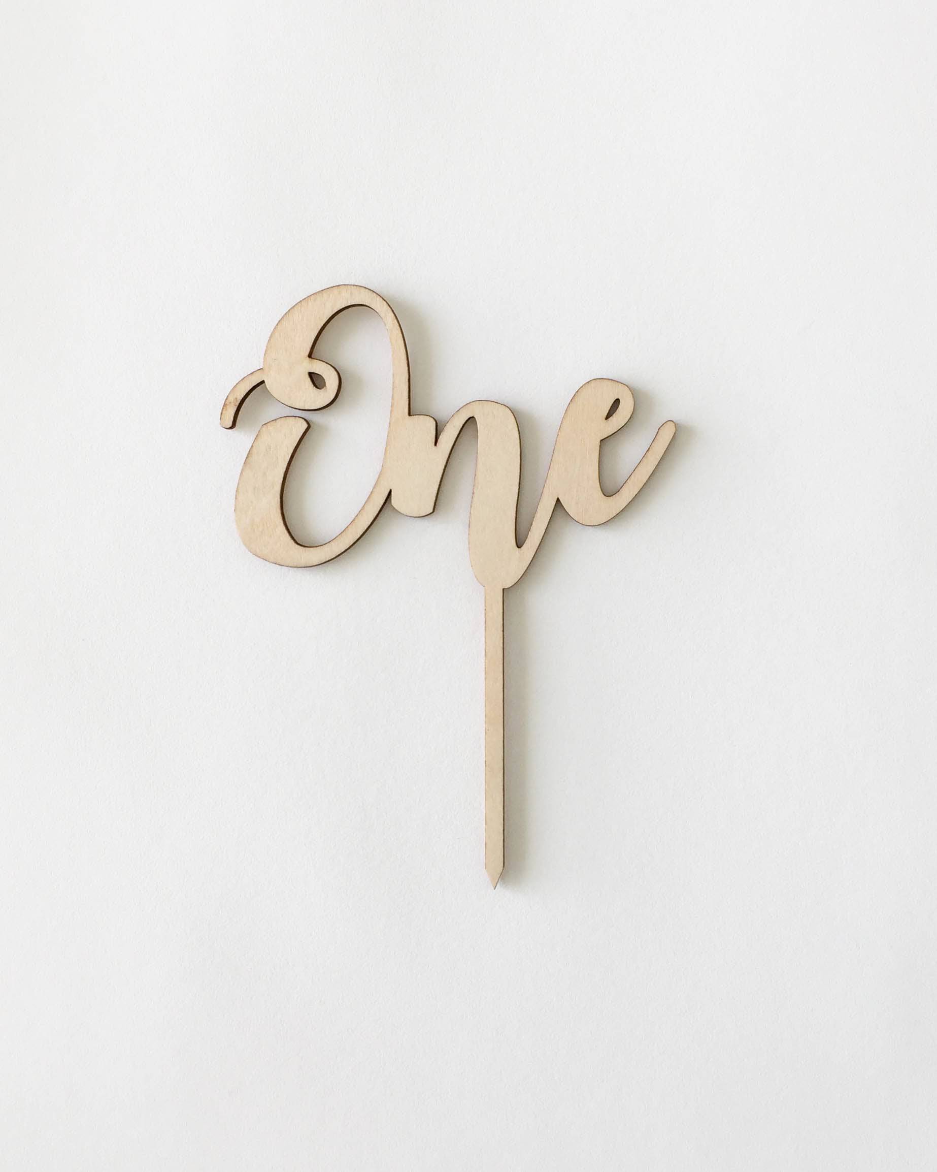 Wooden cake topper featuring the word "One" in an elegant cursive font, set against a light background.