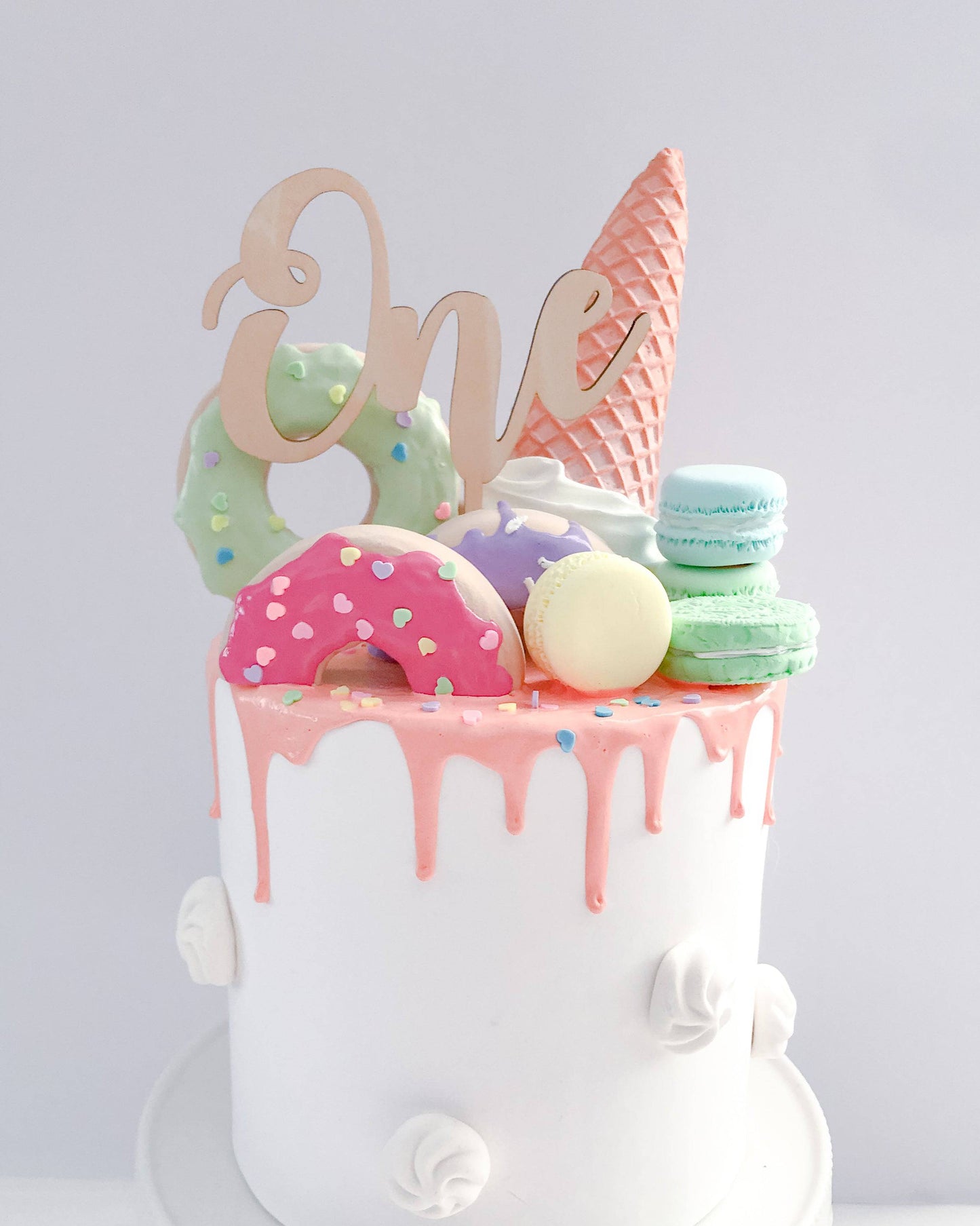 Colorful cake topped with a wooden "One," macarons, donuts, and a cone, featuring pink drip icing and sprinkles.