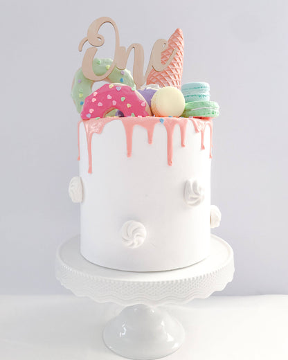 A white cake adorned with colorful sweets and a wooden "One" topper, perfect for a first birthday celebration.