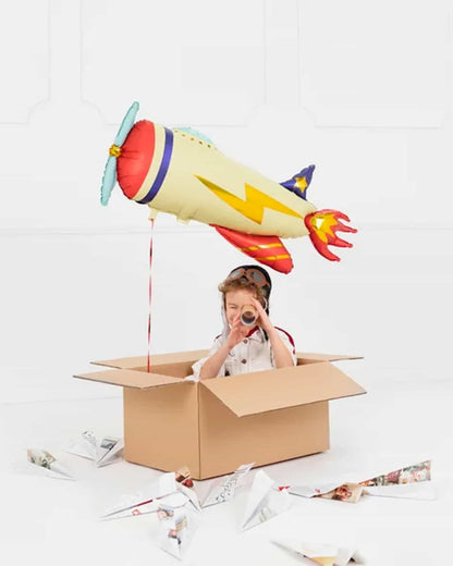 A child in a cardboard box uses a tube as a telescope while a colorful balloon aircraft hovers above. Paper planes scatter ar