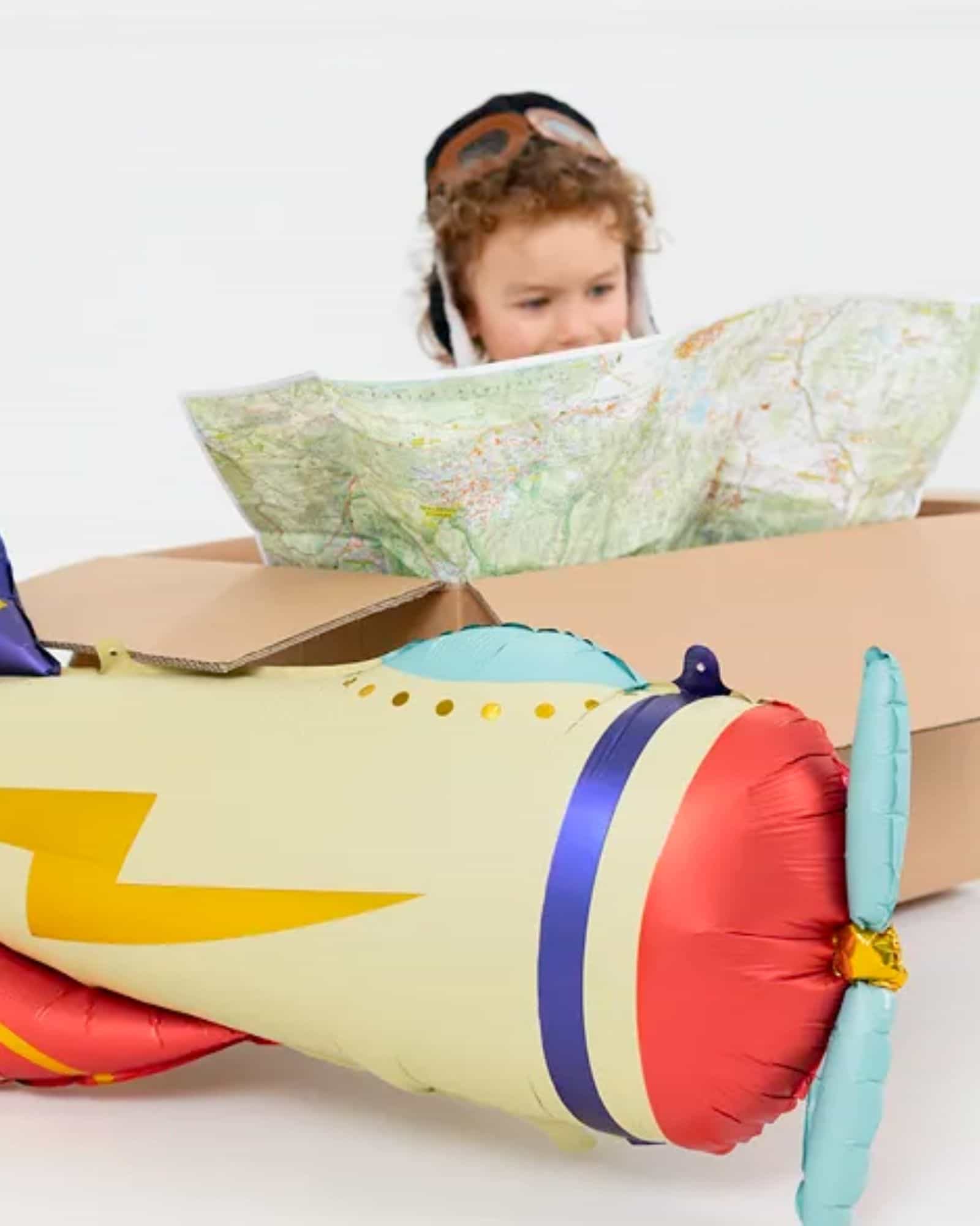 Child in aviator goggles examines a map beside a colorful inflatable airplane and a cardboard box.