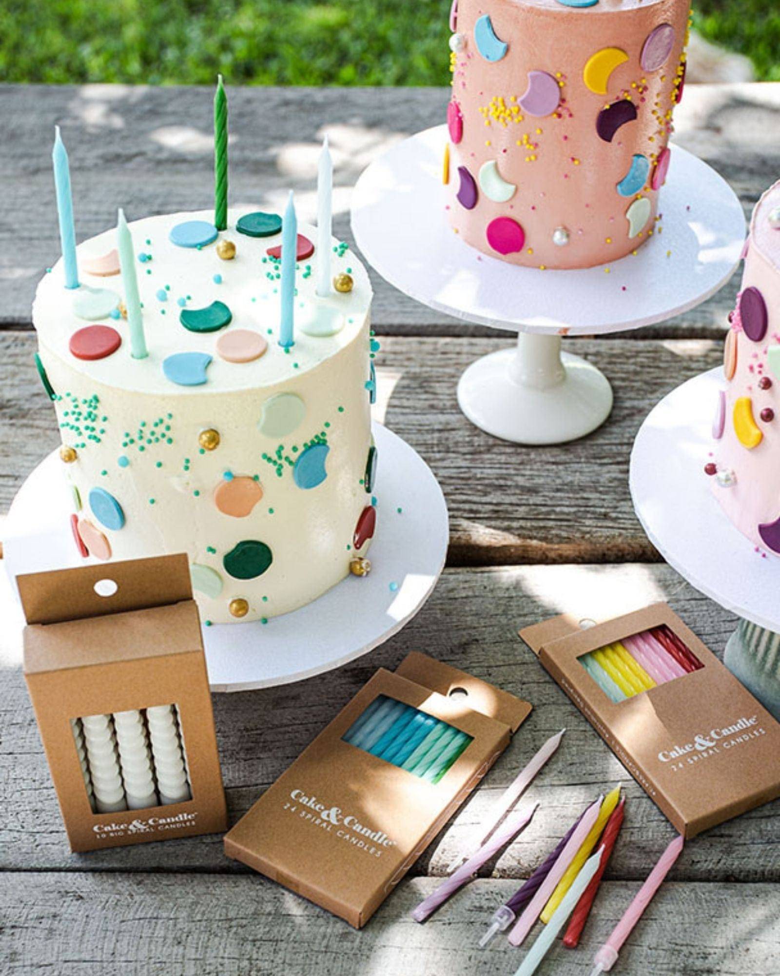 Colorful cakes with decorative dots and candles, surrounded by packaging for additional candles on a rustic wooden table.