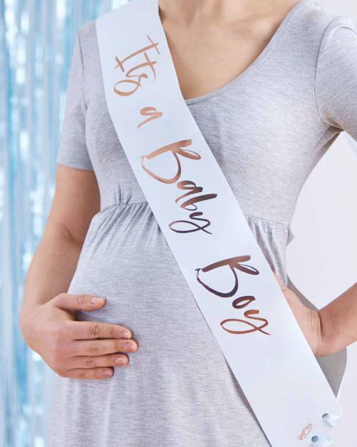 A pregnant person wearing a gray dress and a sash that reads "It's a Baby Boy" with a festive background.