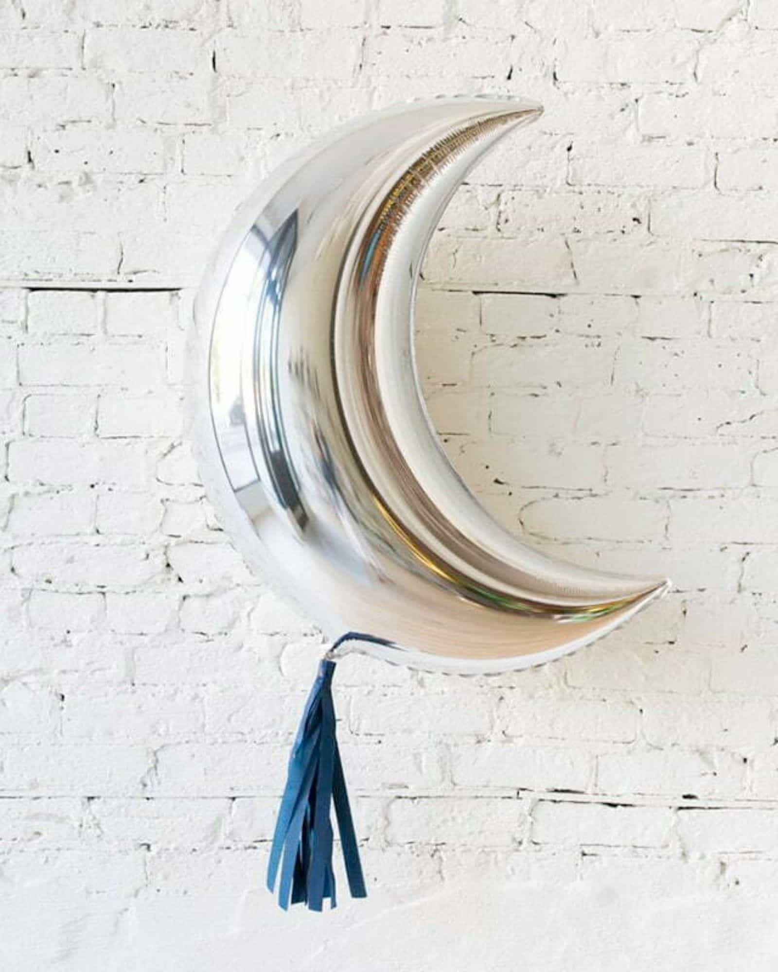 Shiny silver crescent shape with a blue tassel, set against a textured white brick wall.