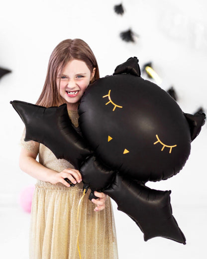 A girl in a sparkly gold dress smiles while holding a large black bat-shaped balloon with cute facial features.