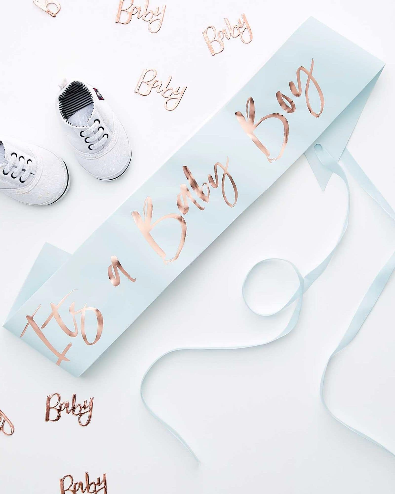 White baby shoes beside a light blue banner stating "It's a Baby Boy" with rose gold lettering and decorative confetti.