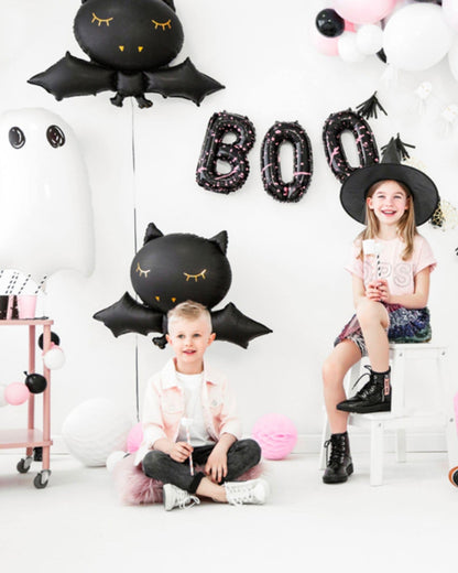 Two children celebrate with playful decorations, including black bat and ghost balloons, and a "BOO" banner.