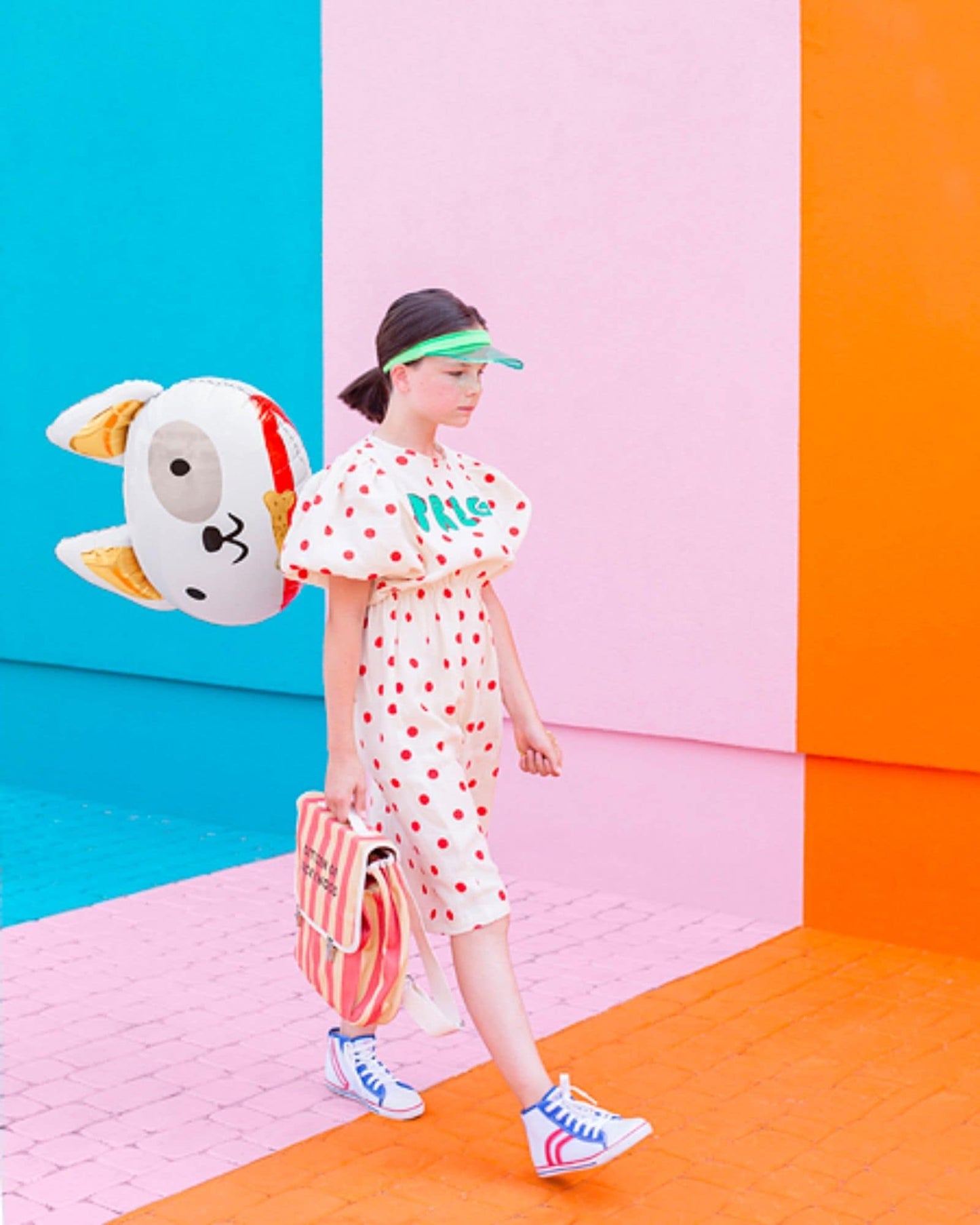 A girl in a polka-dot outfit walks against vibrant colored walls, carrying a striped bag and a whimsical balloon.