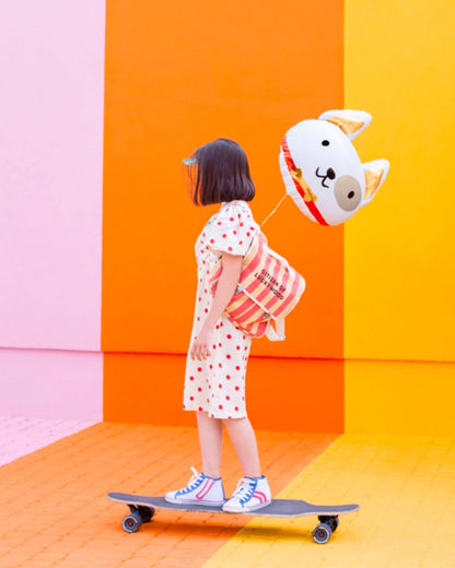 Child with a polka dot dress rides a skateboard, holding a cute animal-shaped balloon against a vibrant backdrop.