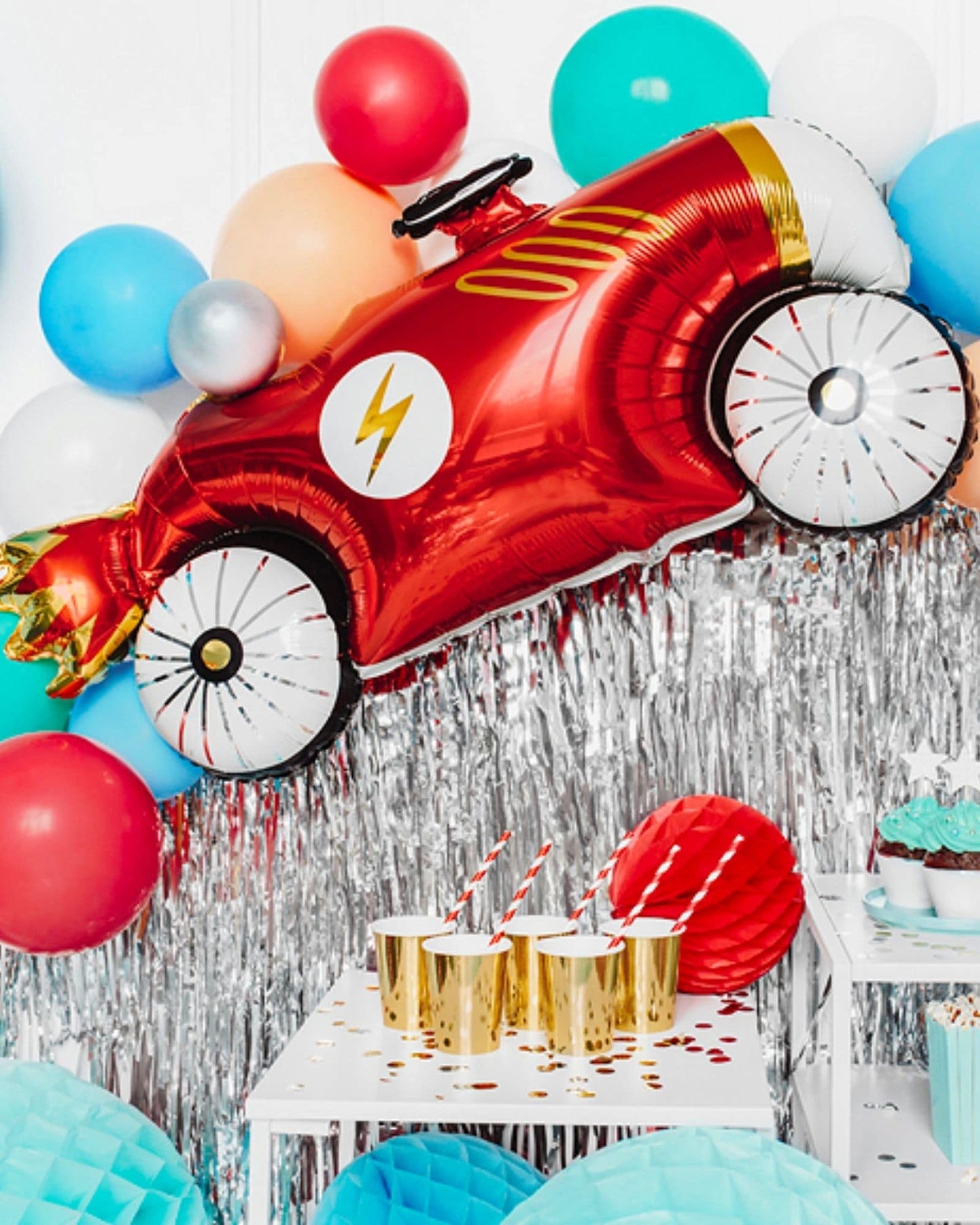 A vibrant red race car balloon surrounded by colorful balloons and shiny decorations, with golden cups in the foreground.