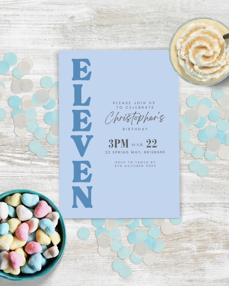 Light blue invitation with bold "ELEVEN," surrounded by colorful heart-shaped candies and confetti on a wooden surface.