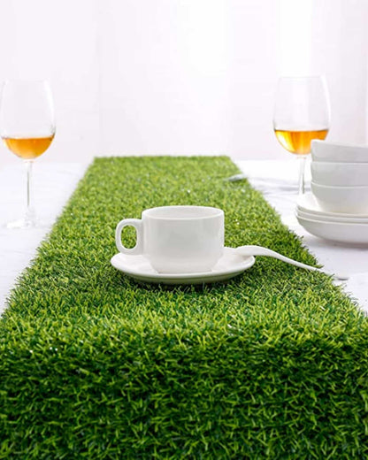 A white cup on a saucer sits on vibrant green grass, with glasses of amber liquid and white dishes in the background.