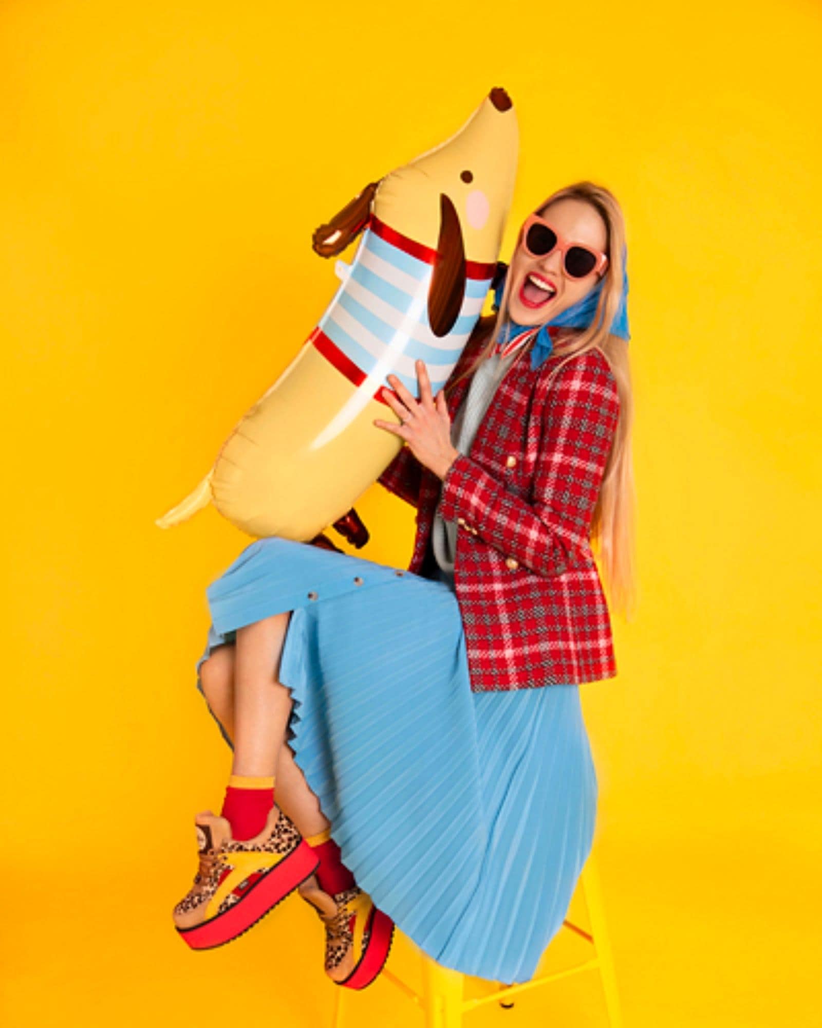 A woman in a plaid blazer and blue skirt joyfully holds a large dog-shaped cushion against a bright yellow backdrop.