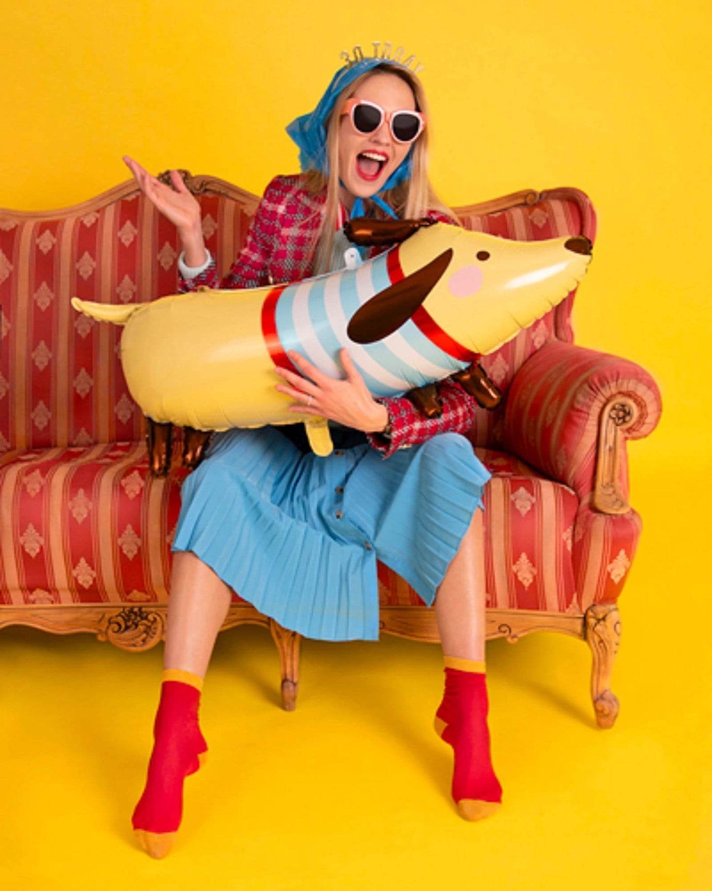 A woman in a colorful outfit sits on a striped sofa, playfully holding a large, cheerful dog-shaped pillow.