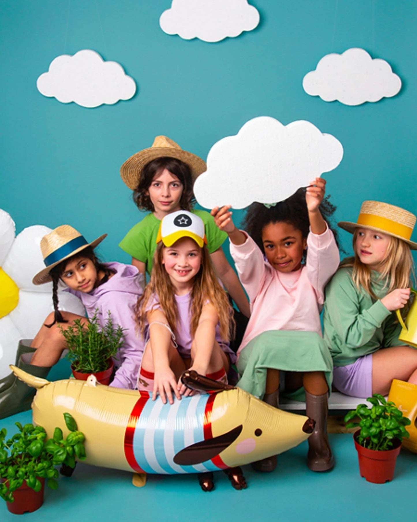 Five children in colorful outfits pose together, surrounded by playful clouds and greenery, holding signs and smiling.