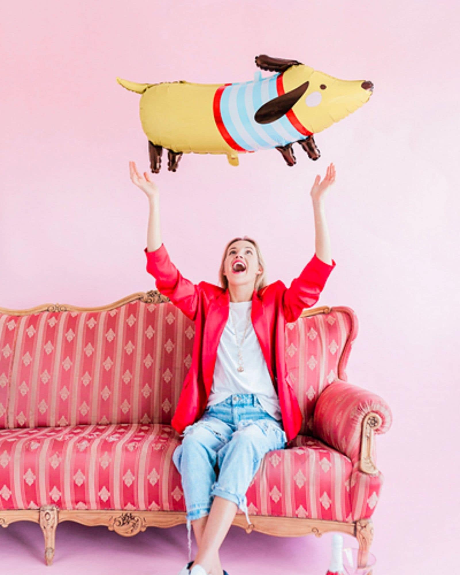 A woman in a bright red jacket joyfully tosses a colorful balloon dog against a playful pink backdrop.