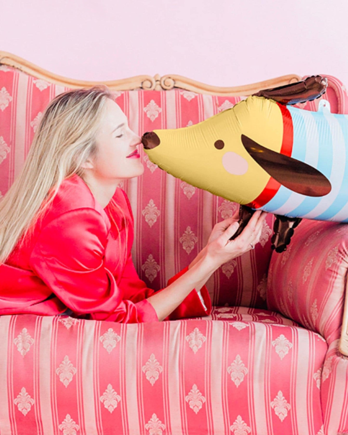 A woman in a red satin outfit playfully interacts with a colorful inflatable dog against a soft pink background.