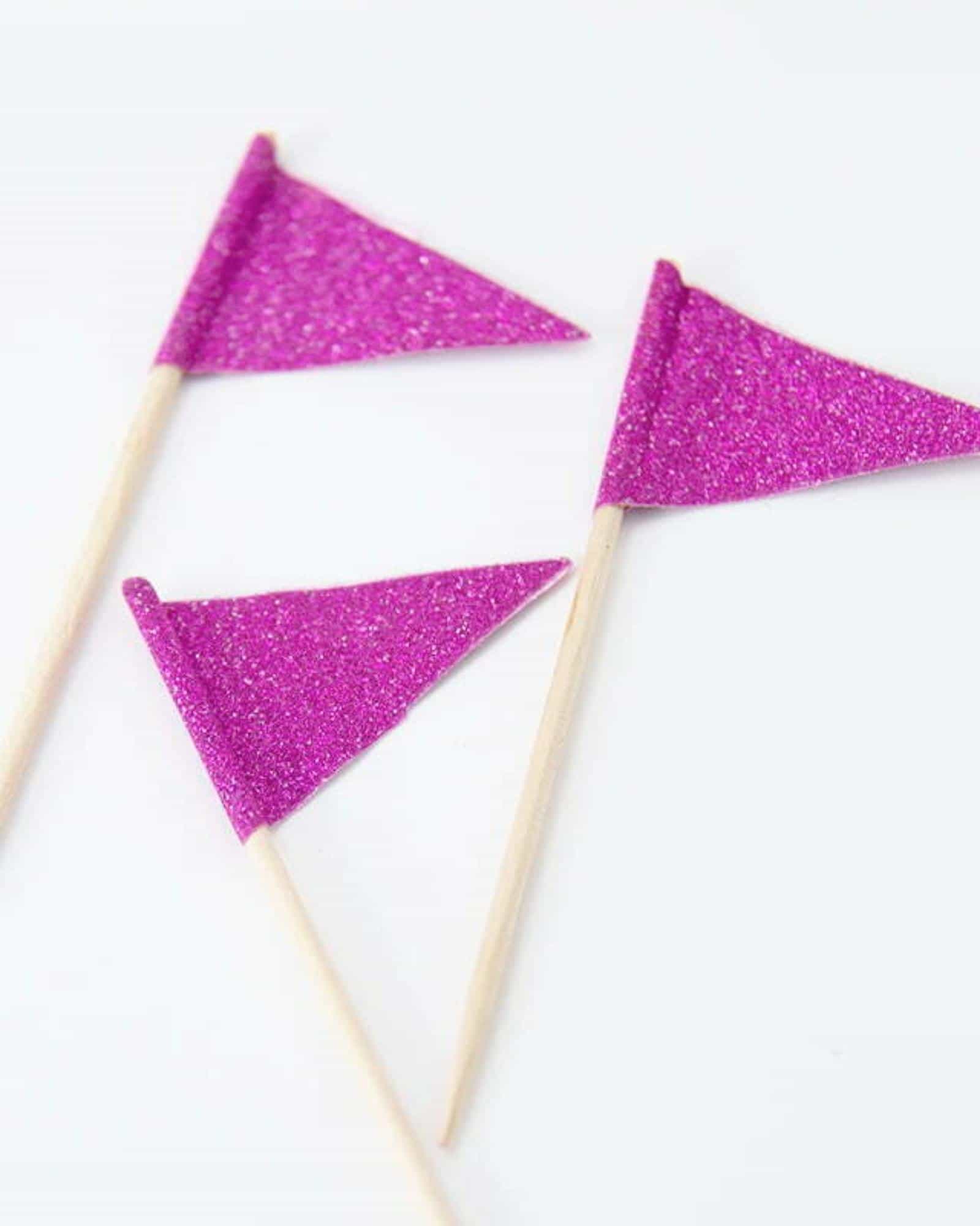 Three small, glittery purple flags perched on wooden sticks against a white background, adding a festive touch.