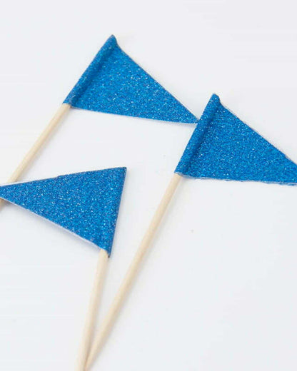 Three small blue glitter flags on wooden sticks resting on a clean, white surface.