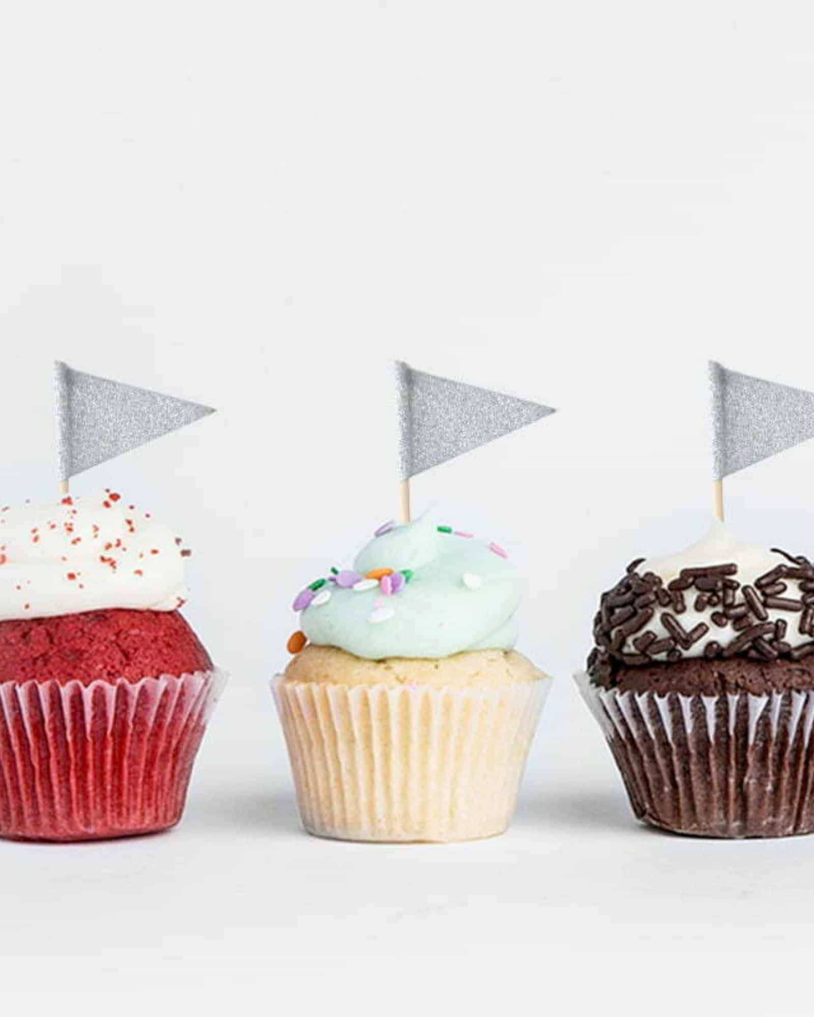 Three colorful cupcakes topped with frosting and decorative flags, arranged on a white background.