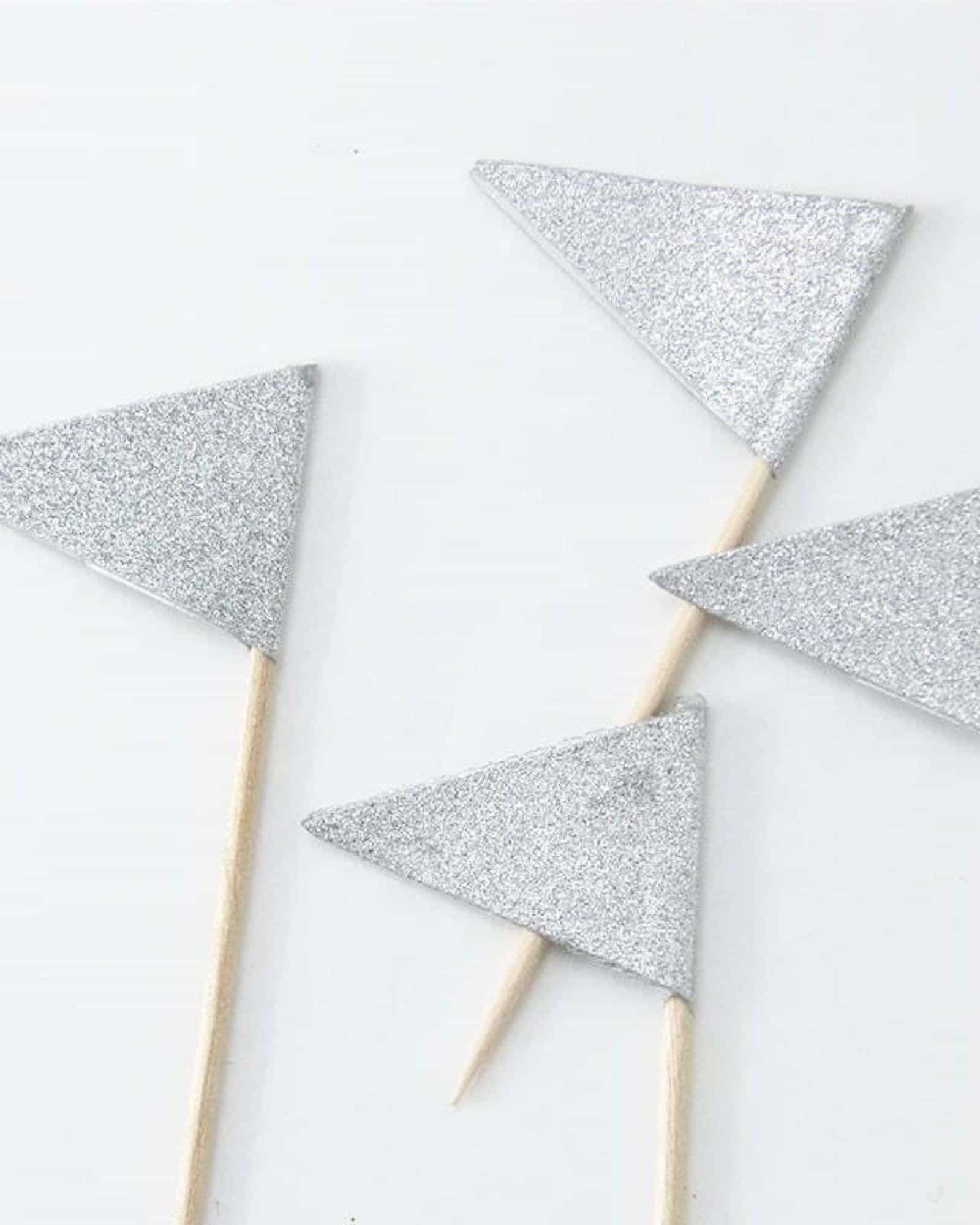 Four small silver glitter triangular shapes attached to wooden sticks on a light background.