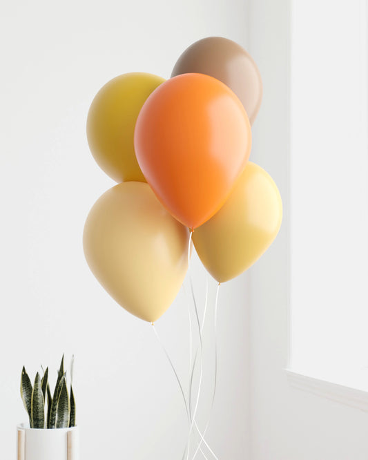 A cluster of warm-toned balloons in orange, yellow, and brown, gently swaying beside a green plant in a bright room.