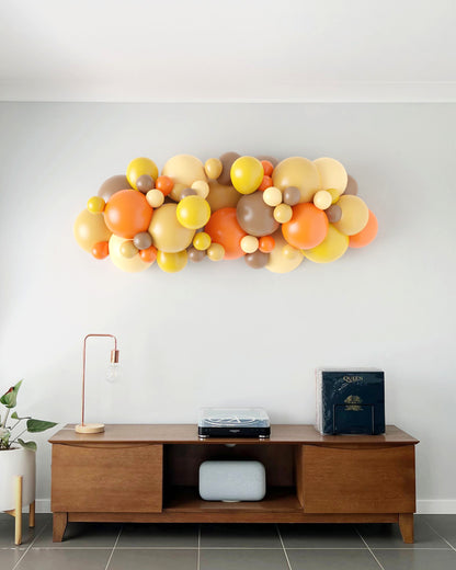 A vibrant balloon garland in warm autumn colors decorates a wall above a wooden sideboard with a lamp and plants.