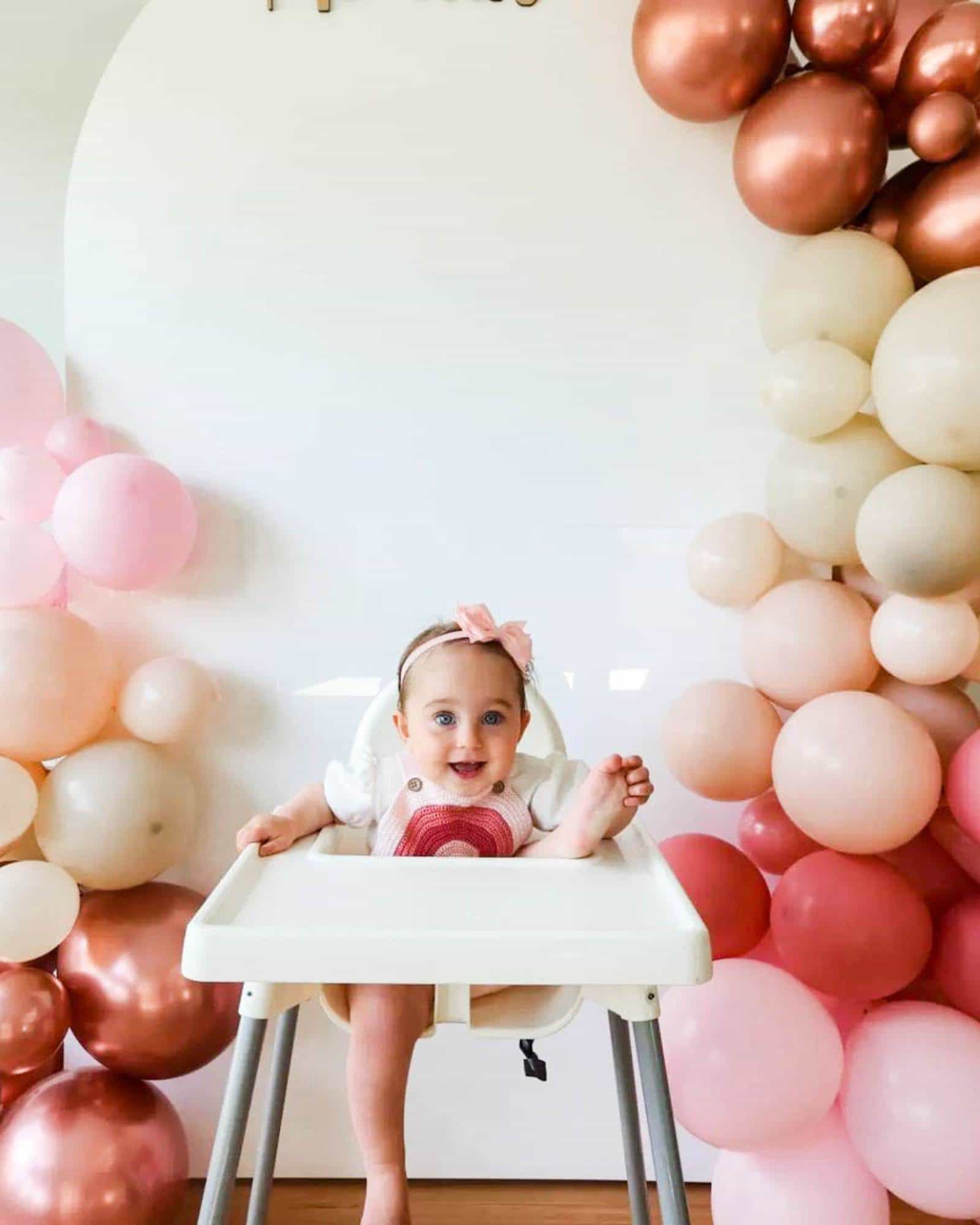 A smiling baby in a high chair surrounded by pastel and metallic balloons, with a bright, cheerful backdrop.