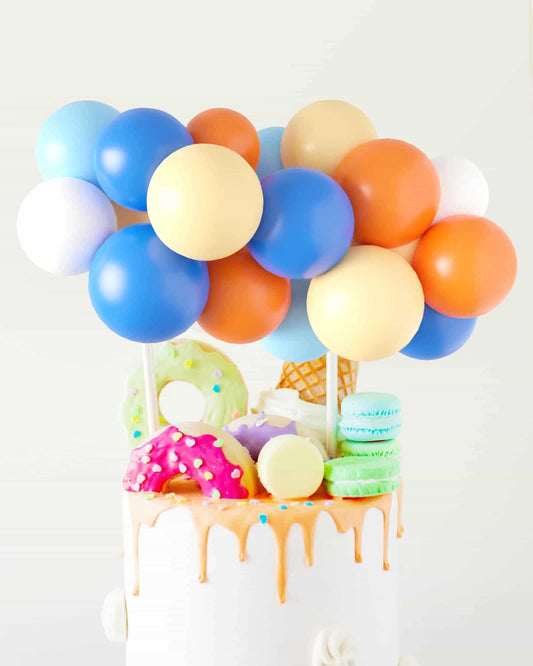 Colorful balloons hover over a cake topped with vibrant donuts, ice cream cones, and pastel macarons.