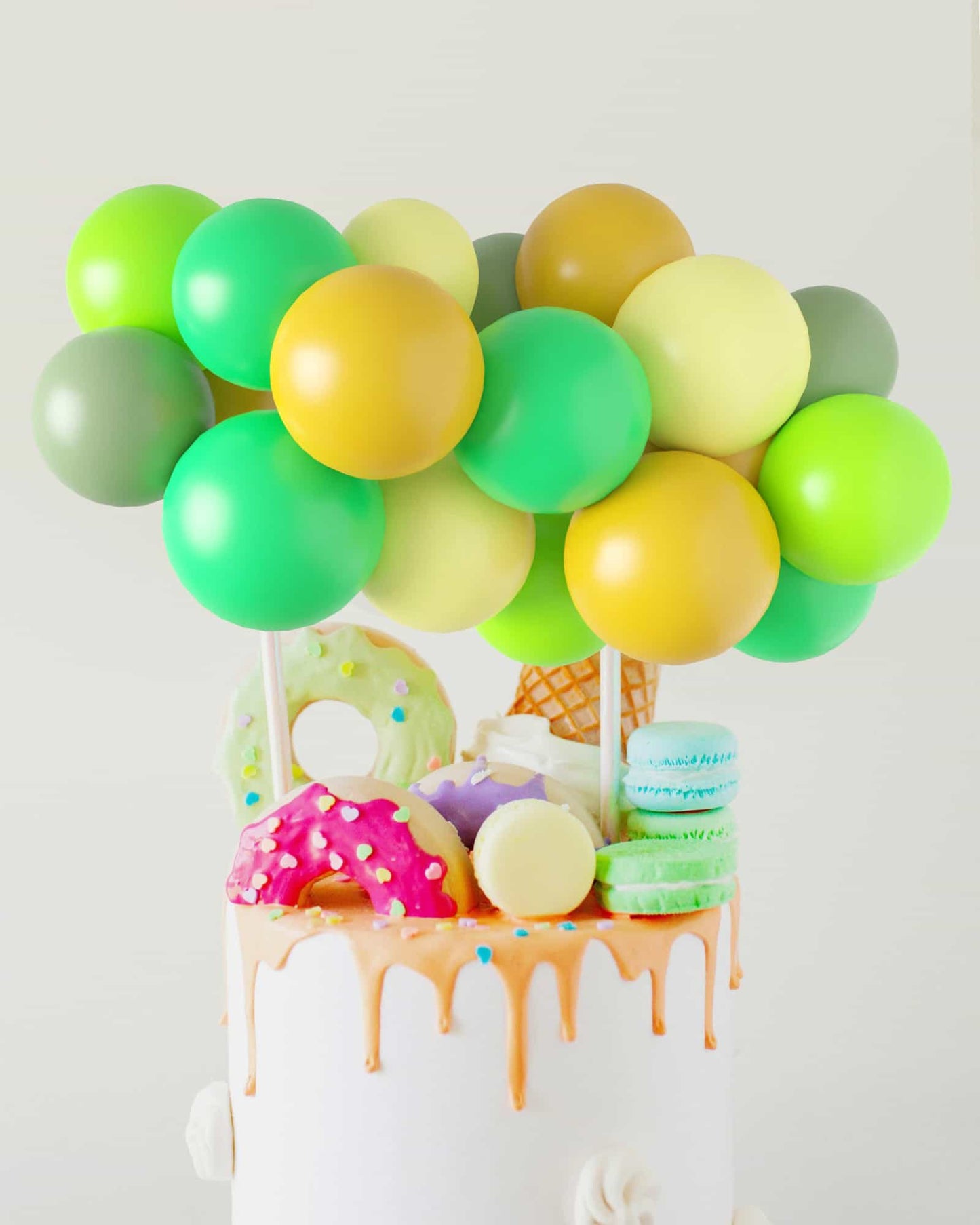 Colorful balloons in shades of green and yellow hover above a decorated cake topped with vibrant sweets and treats.