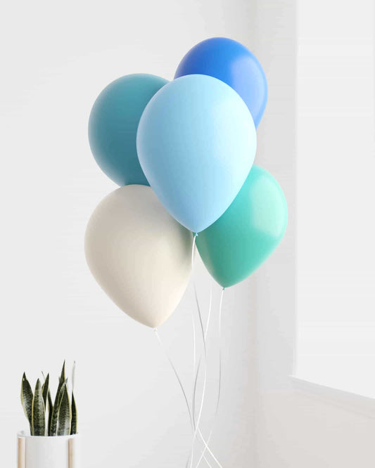 A cluster of colorful balloons in shades of blue and green, with a white base, beside a potted plant against a bright wall.