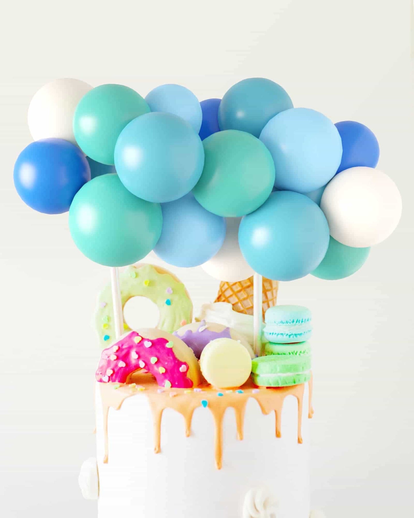 Colorful balloons in shades of blue and green atop a cake adorned with pastel treats and dripping icing.