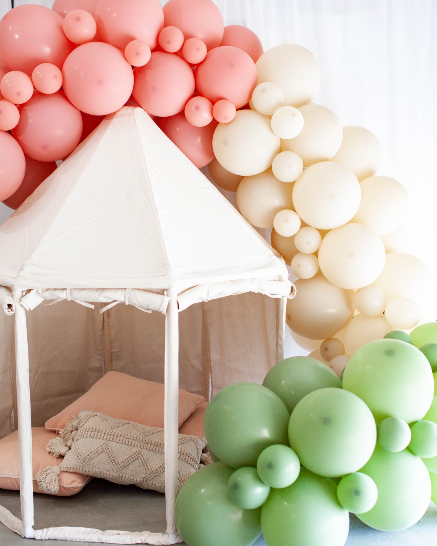 A soft play tent surrounded by pastel balloons in pink, cream, and green, with cozy cushions inside.